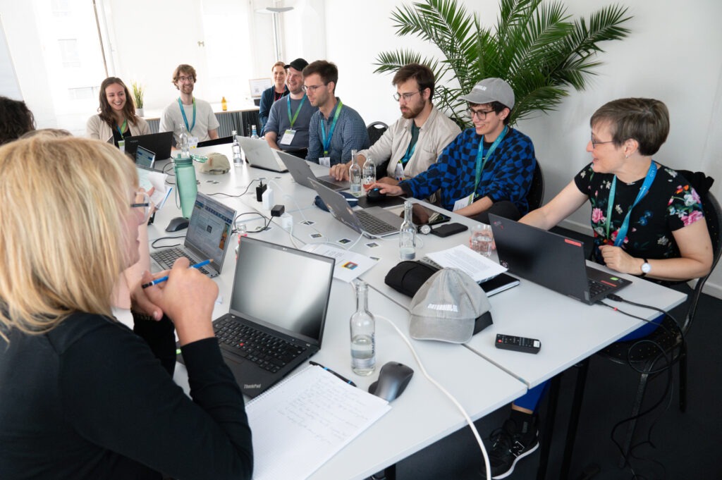 Teammitglieder und Teilnehmer:innen eines Workshops von CorrelAid sitzen um einen großen Arbeitstisch herum und schauen teilweise in ihre Laptops