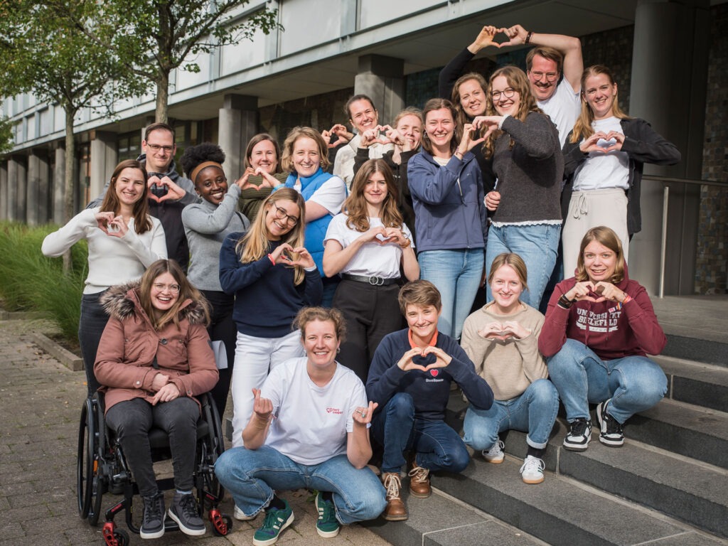 Das Team der Organisation Herzkaspar e.V. stehen auf der Treppe vor einem Gebäude und bilden mit ihren Händen ein Herz